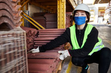 Bir araç olarak işinin ortasında maske, eldiven ve kask takan genç bir kadın ve beton çatı kiremitleri alan malzeme satıcısı, yeni normallik döneminde, covid 19 salgınının ortasında