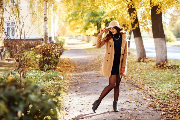 Стильная женщина в широкополой шляпе и бежевом пальто проводит время на свежем воздухе осенью — стоковое фото
