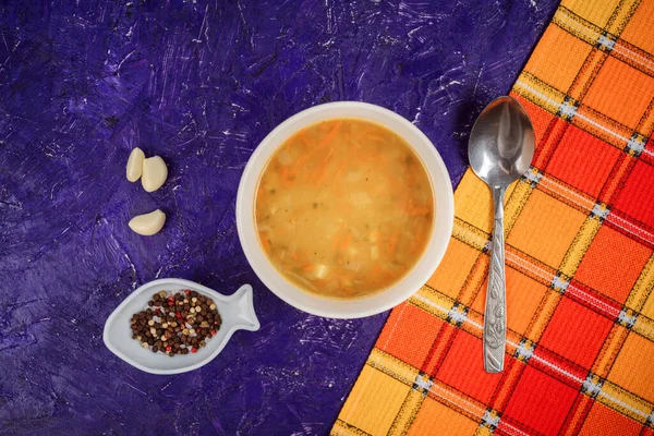 Вкусный морковный суп, чеснок, перец, ложка на фиолетовом фоне и оранжевое полотенце . — стоковое фото