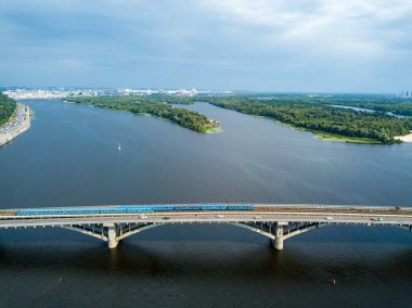 Kiev metrosunun trenleri Dinyeper nehri üzerindeki köprüye gidiyor. Güneşli bir yaz günü. Hava aracı görünümü.