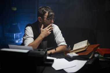 Elinde kağıt, gözlük, klasik bir daktilo ve sigarayla masada oturan genç bir yazarın yakın plan portresi. Ev ödevi, serbest çalışma, çevrimiçi öğrenme, konsept öğrenme. Zorluklar ve sorunlar