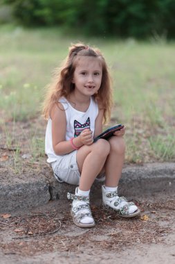 İki ön dişi olmadan beyaz dişlerini gösteren gülümseyen güzel sarışın beyaz kızın portresini yakından çek. Kaldırım kenarına oturur ve telefonu tutar. Çocukluk