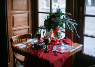 Pencere kenarındaki bir restoranda iki kişilik düğün masası. Düğün dekoru - şeffaf tabaklar, şaraplı bardaklar, saten kırmızı kurdeleli güzel bir düğün buketi, tahta sandalyeler ve arka plan.