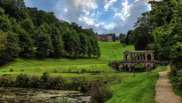 Mystical landscape garden, Palladian bridge, and ancient architecture. River, trees, and blue sky with clouds.