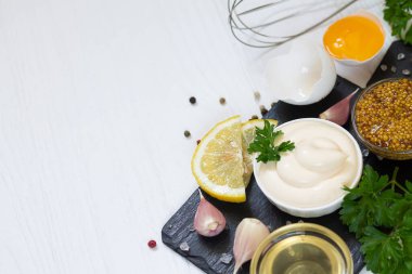Beyaz arka planda siyah taş tahtada pişirmek için kullanılan malzemelerin yanına bir kase mayonez sosu. 
