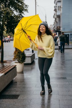 Büyüleyici genç kıvırcık kadın yağmurlu günlerde Megapolis caddesinde sarı şemsiye kullanıyor.