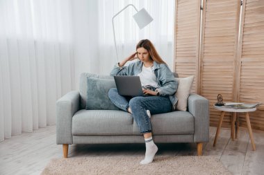 Öğrenci genç kız evdeki kanepede uzanıyor, dizüstü bilgisayara bakıyor ve Webinar 'da rol alıyor yeni bilgiler ediniyor ya da arkadaşlarıyla uzaktan iletişim kuruyor.