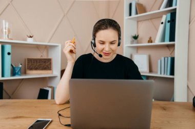 Gülümseyen mutlu kulaklıklı iş kadını masaya oturur, dizüstü bilgisayara bakar, notlar alır, kişisel gelişim webinarına katılır, internette eğlenir, video aracılığıyla iletişim kurar.