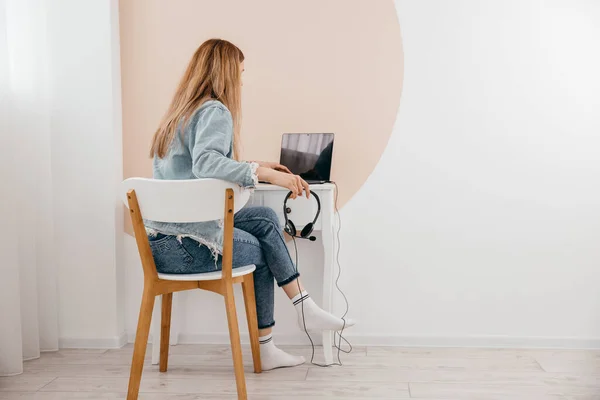 Evde dizüstü bilgisayar ve kulaklıklarla masada oturan ve uzaktan internetten çalışan serbest çalışan genç bir kadın. Konsantre genç kız oturma odasında uzaktan tek başına ders çalışıyor..