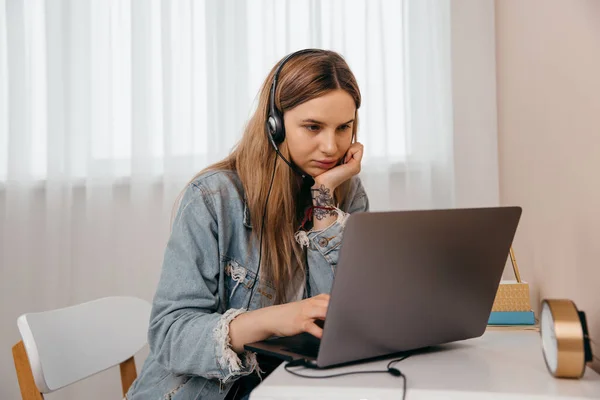 Evde dizüstü bilgisayar ve kulaklıklarla masada oturan ve uzaktan internetten çalışan serbest çalışan genç bir kadın. Konsantre genç kız oturma odasında uzaktan tek başına ders çalışıyor..