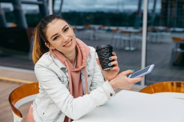 Gülümseyen genç kadın, alışveriş yaptıktan ve kahve fincanı tuttuktan sonra taksiye binmek için akıllı telefon kullanıyor. Kentin arka planında uzun beyaz ekmek ekmeği olan alışveriş poşeti.