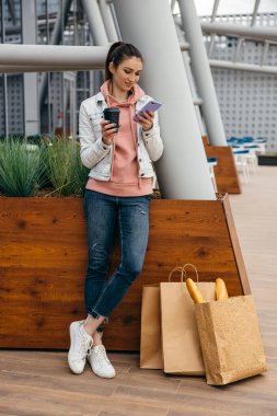 Gülümseyen genç kadın, alışveriş yaptıktan ve kahve fincanı tuttuktan sonra taksiye binmek için akıllı telefon kullanıyor. Kentin arka planında uzun beyaz ekmek ekmeği olan alışveriş poşeti.