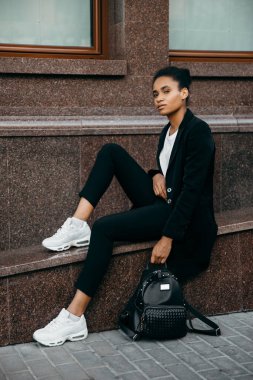 Genç, şık, afro-amerikan iş kadını ceketli, şehirde merdivenlerde oturuyor ve bir çanta tutuyor..