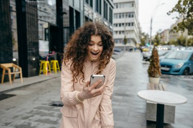 Güzel gülüşlü, çekici, genç, kıvırcık kadın, şehrin sokaklarında kal ve akıllı telefon kullan ve şaşırt.