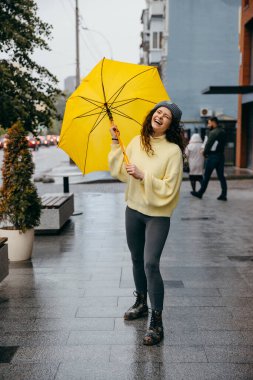 Büyüleyici genç kıvırcık kadın yağmurlu günlerde Megapolis caddesinde sarı şemsiye kullanıyor.