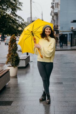 Büyüleyici genç kıvırcık kadın yağmurlu günlerde Megapolis caddesinde sarı şemsiye kullanıyor.