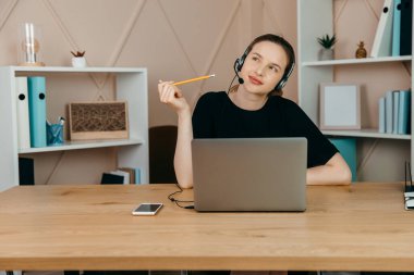 Gülümseyen mutlu kulaklıklı iş kadını masaya oturur, dizüstü bilgisayara bakar, notlar alır, kişisel gelişim webinarına katılır, internette eğlenir, video aracılığıyla iletişim kurar.