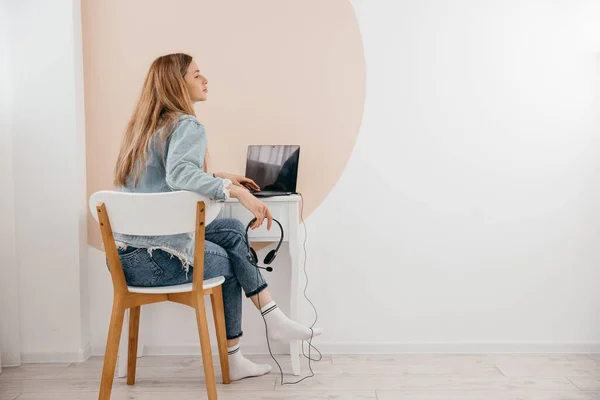 Evde dizüstü bilgisayar ve kulaklıklarla masada oturan ve uzaktan internetten çalışan serbest çalışan genç bir kadın. Konsantre genç kız oturma odasında uzaktan tek başına ders çalışıyor..