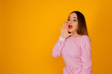 Sarı arka planda izole edilmiş zarif pembe tişörtlü utangaç genç kadın portresi. Çığlık atıyor.