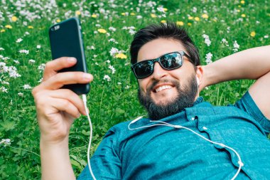 Parkta yeşil çim üzerinde yalan, müzik dinlemek ve smartphone kullanarak yakışıklı adam