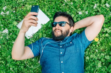 Parkta yeşil çim üzerinde yalan, müzik dinlemek ve smartphone kullanarak yakışıklı adam