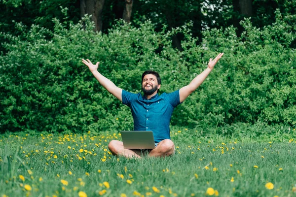 微笑的年轻人坐在绿色的草地上举起双手 用笔记本电脑在他交叉的腿上 — 图库照片