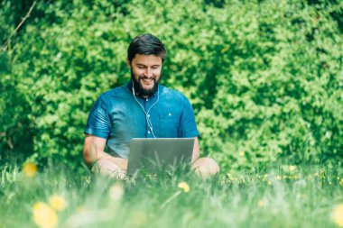 Bilgisayarda çalışarak ve çim üzerinde oturan genç modern işadamı serbest meslek. Erkek öğrenci Not defteri açık havada kullanarak. Dizüstü bilgisayarda çalışan Turizm