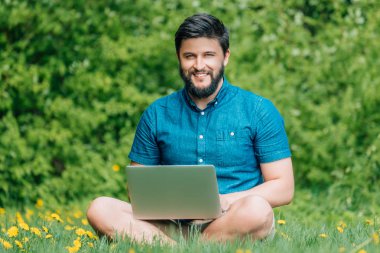 Bilgisayarda çalışarak ve çim üzerinde oturan genç modern işadamı serbest meslek. Erkek öğrenci Not defteri açık havada kullanarak. Dizüstü bilgisayarda çalışan Turizm