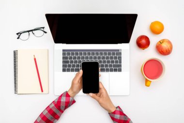 Kadın bilgisayar ve malzemelerle beyaz ofis masasının üzerinde boş ekranlı akıllı telefon kullanıyor. Üst Manzara. Düz yatıyordu