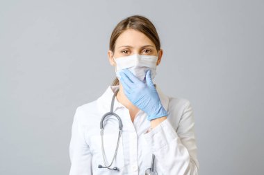 Gri arka plana steril maske takan genç bir kadın doktor ya da hemşire portresi.