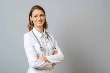 Kolları gri arka planda çaprazlanmış laboratuvar önlüklü gülümseyen kadın doktor.