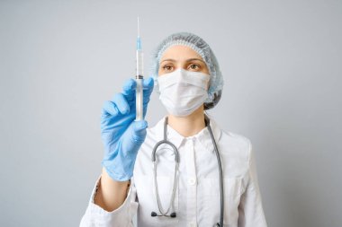 Tıbbi üniformalı, koruyucu maskeli ve elinde şırınga olan genç bir kadının portresi. Gri arkaplanda izole