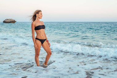 Akşamları kumsalda siyah bikinili genç bir kadın. Arka planda deniz ve kaya var.