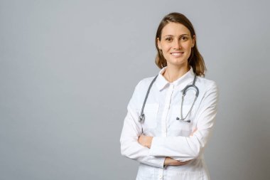Kolları gri arka planda çaprazlanmış laboratuvar önlüklü gülümseyen kadın doktor.