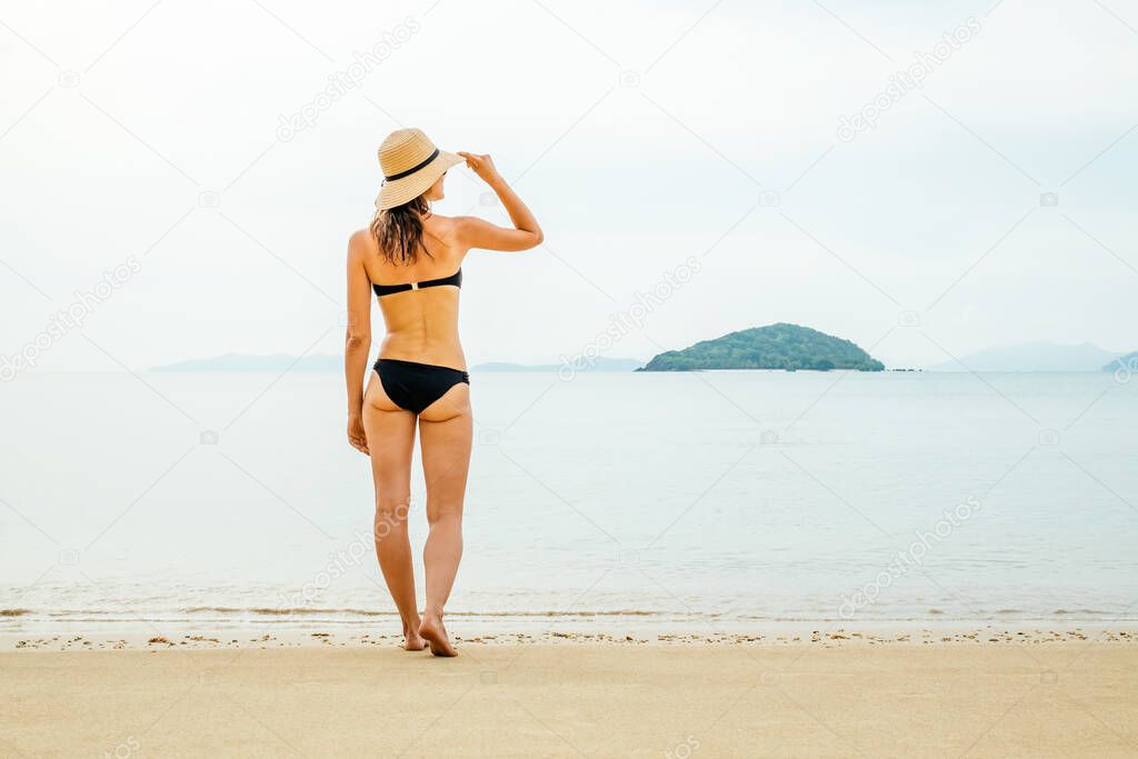 Beach vacation. Beautiful woman in sunhat and bikini standing at the beach enjoying looking view of ocean on summer day