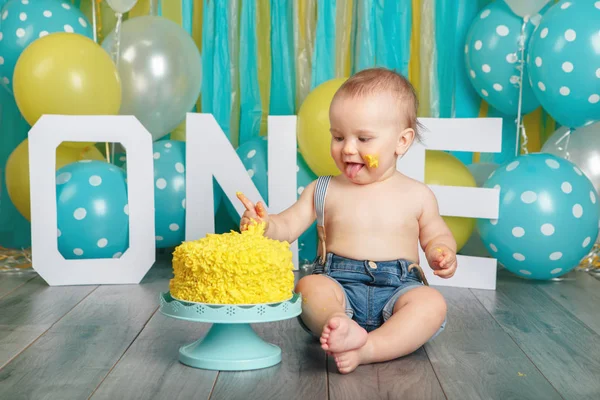 Portrait of cute adorable Caucasian baby boy in jeans pants celebrating his first birthday. Cake smash concept. Child kid sitting on floor in studio eating tasty yellow dessert