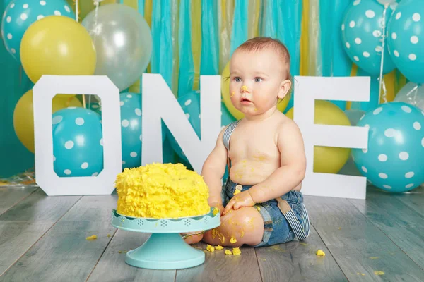 Portrait of cute adorable Caucasian baby boy in jeans pants celebrating his first birthday. Cake smash concept. Child kid sitting on floor in studio eating tasty yellow dessert