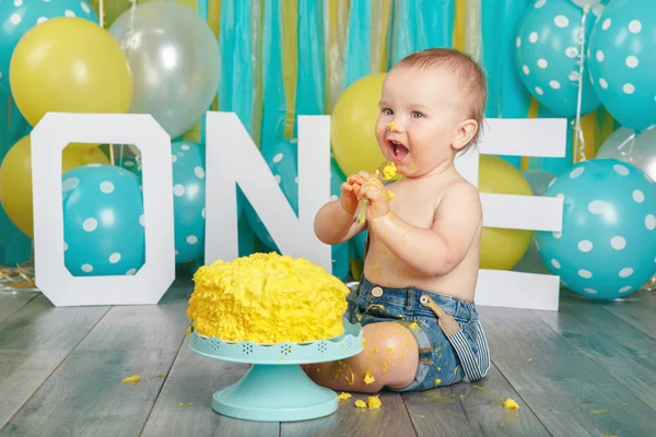 Portrait of cute adorable Caucasian baby boy in jeans pants celebrating his first birthday. Cake smash concept. Child kid sitting on floor in studio eating tasty yellow dessert with spoon