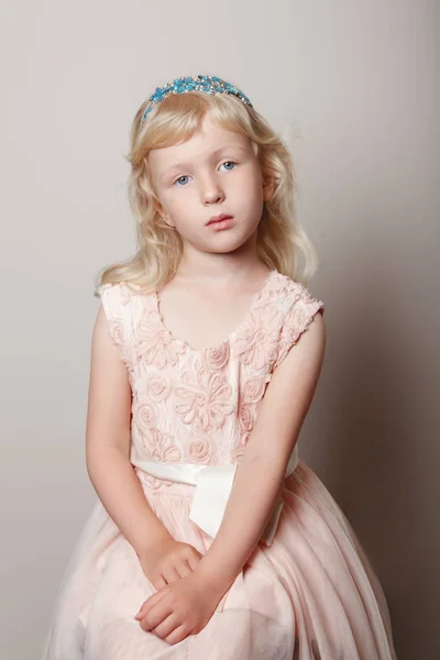 Closeup portrait of cute adorable white blonde fair Caucasian preschool girl in pink dress and princess crown. Pensive sad hild posing in studio on plain light background.