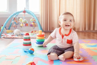 Portrait of cute adorable blond Caucasian smiling child boy with blue eyes sitting on floor in kids children room. Little baby playing with toys on playmat at home. Early education development concept. clipart