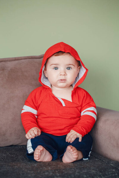 Portrait of cute adorable Caucasian smiling baby boy with blue eyes, wearing red sport hoodie shirt and tracksuit pants. Seven months child sitting on couch at home looking in camera