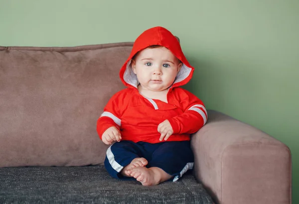 肖像可爱可爱的白种人微笑的小男孩蓝眼睛 穿着红色运动衫帽衫和运动服裤 七月的孩子坐在沙发上在家里看着相机 — 图库照片