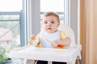 Sevimli sevimli beyaz çocuk çocuk sandalyesi yeme elma püresi kaşıkla oturan kirli pis yüzü ile portresi. Her gün eve çocukluk bir yaşam tarzı. Bebek çalışırken takıma giren bebek maması