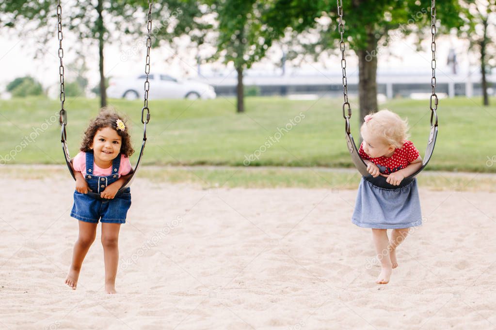 Portrait of two happy smiling little toddlers girls friends swinging on swings at playground outside on summer day. Happy childhood lifestyle concept. Toned with film pastel faded filters colors.