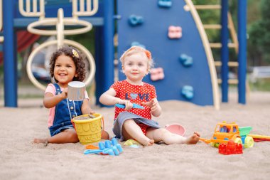 İyi günler. İki sevimli beyaz ve İspanyol latin plastik renkli oyuncaklar ile oynama sandbox oturan çocuk bebekler. Küçük kız arkadaş sahada birlikte eğleniyor.