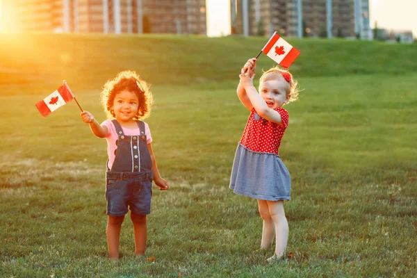 Tutuşup gülümseyen ve park açık havada içinde Kanada bayrakları sallayarak mutlu sevimli küçük sarışın beyaz ve İspanyol latin girsl. Çok ırklı çocuklar Kanada günü kutluyor. Bayrak günü kavramı.