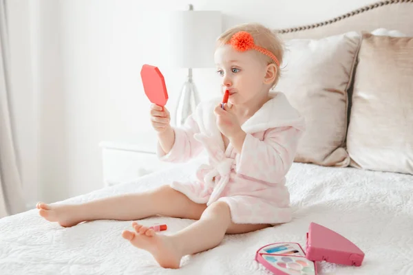 可爱的 Asorable 滑稽的白种金发女孩2岁 穿着浴衣做化妆 看着镜子 坐在她的床上在家里的卧室 快乐童年概念 — 图库照片