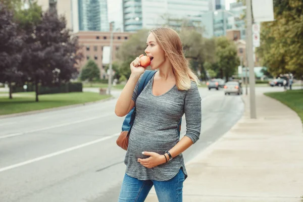 Happy healthy pregnancy. Portrait of caucasian pregnant young blonde Caucasian woman eating apple . Beautiful slim sporty expecting mom lady in busy urban city outside.