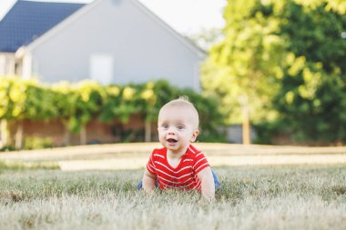 Sevimli sevimli küçük Kafkas oğlu kırmızı tişört ve pantolon alan çayır dışında oturuyor. Günbatımı yaz Park'ta küçük mutlu gülen çocuk. Güzel çekici erkek bebek eğleniyor.