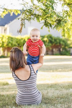 Group portrait of Caucasian mother with baby boy in red t-shirt in field meadow outside. Female parent holding little happy smiling son child in summer park on sunset. Happy parenting lifestyle.